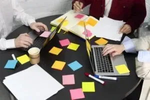 Three people collaborate at a round table with laptops and colorful sticky notes. Coffee cups and a marker are scattered about as they leverage virtual assistant tools to boost productivity in their creative process.