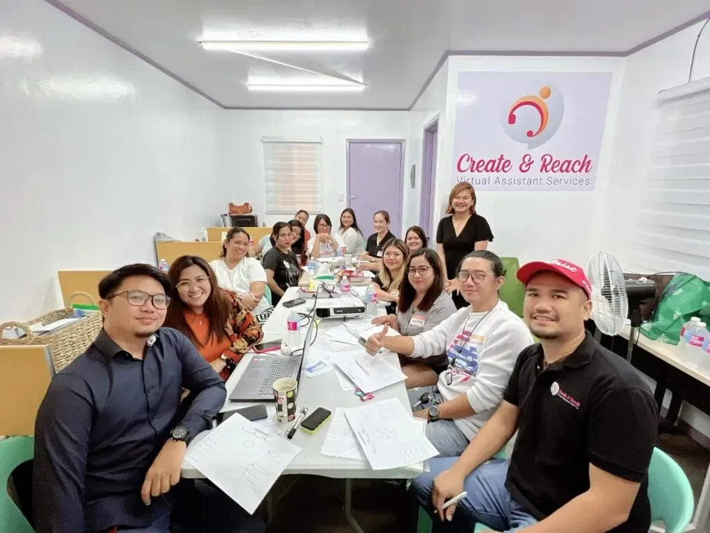 A group of people gathers around a table in an office, sifting through paperwork and using a laptop. A sign on the wall reads