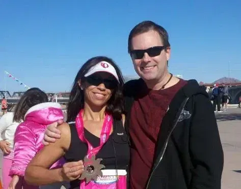 A woman holding a medal stands next to a man. Both are wearing sunglasses and smiling, exuding joy like creating and reaching new milestones. The background shows blue sky and a few people.