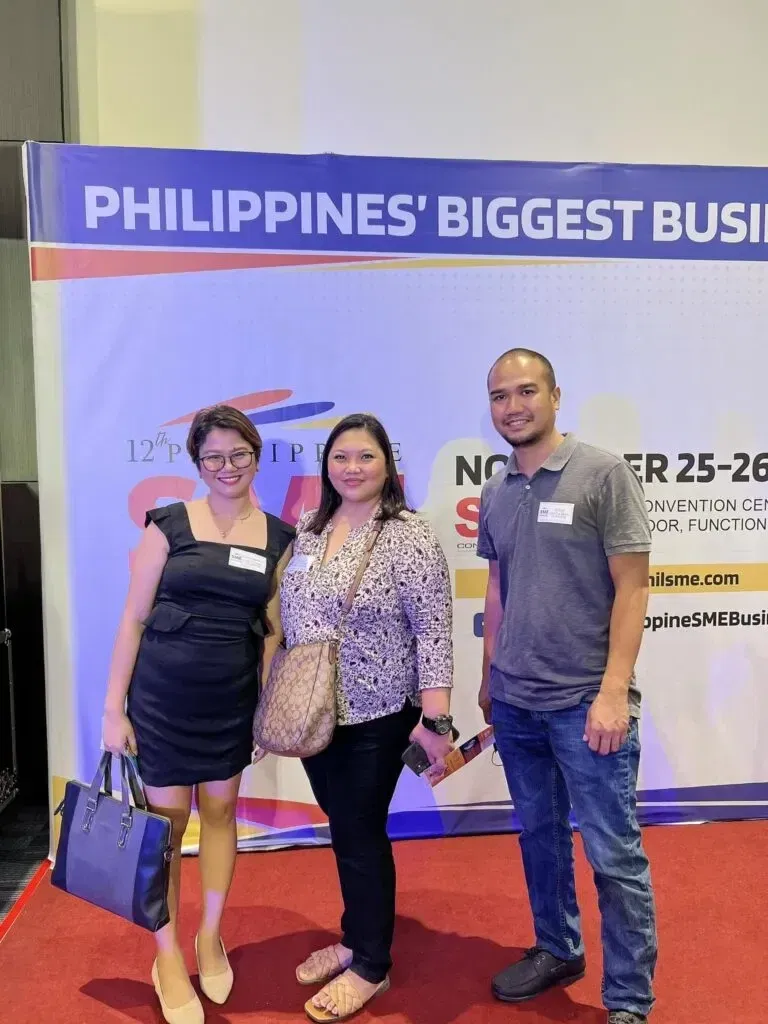 Three people are standing on a red carpet in front of a banner that reads