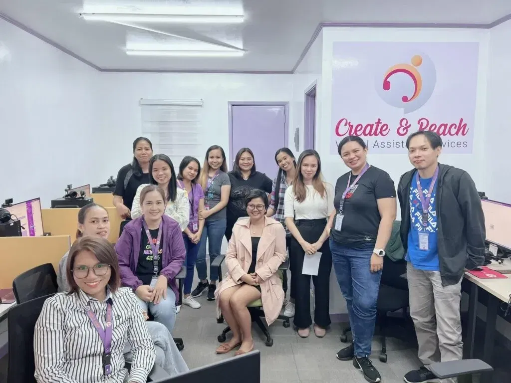 A group of people in an office setting, some seated and some standing, smiling at the camera. A sign on the wall reads