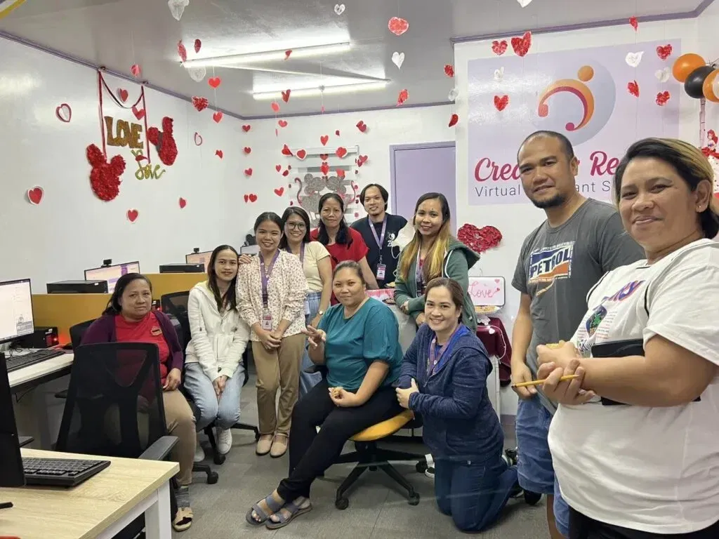 A group of people in an office decorated with red heart ornaments pose for a photo, showcasing the vibrant and collaborative spirit of Create and Reach VA. Computers are on desks, and a sign on the wall proudly displays