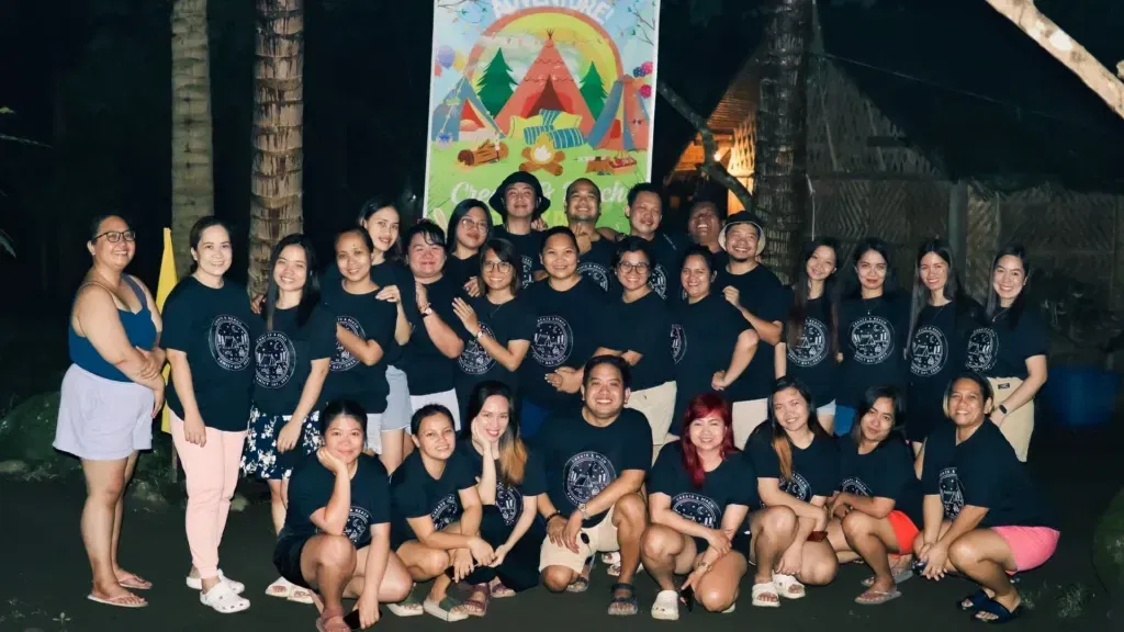 A group of people wearing matching shirts from Create and Reach VA poses in front of a colorful sign at night, showcasing the vibrant spirit of the Virtual Assistant Philippines community.