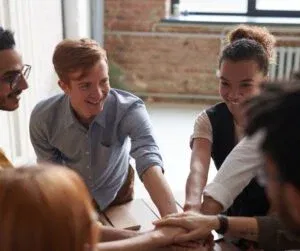 A group of people sitting together in a circle, placing their hands in the center, showcasing teamwork and collaboration crucial for client retention.