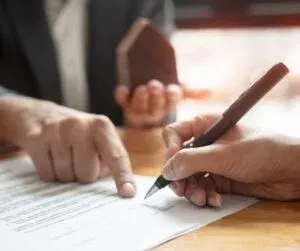 Two people at a table, one pointing at a document while the other signs it. A small house model is held by one individual, symbolizing business security as they finalize the non-compete agreement.