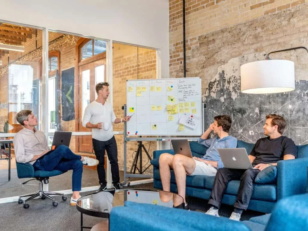 Four people in a casual office setting, three seated with laptops and one standing by a whiteboard, engaged in discussion.