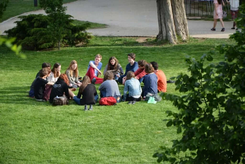 A group of people sitting in a circle on a grassy area in a park, engaged in conversation.