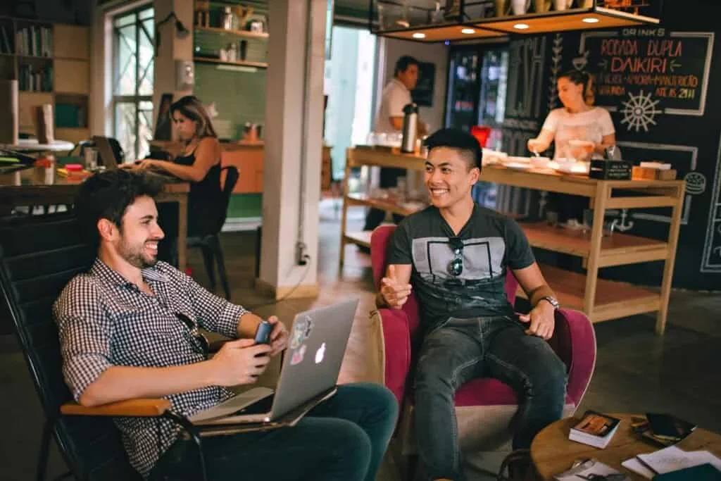 Two men sit and smile at each other in a casual cafe setting, one with a laptop, the other holding a phone. A woman works and another uses the counter in the background.