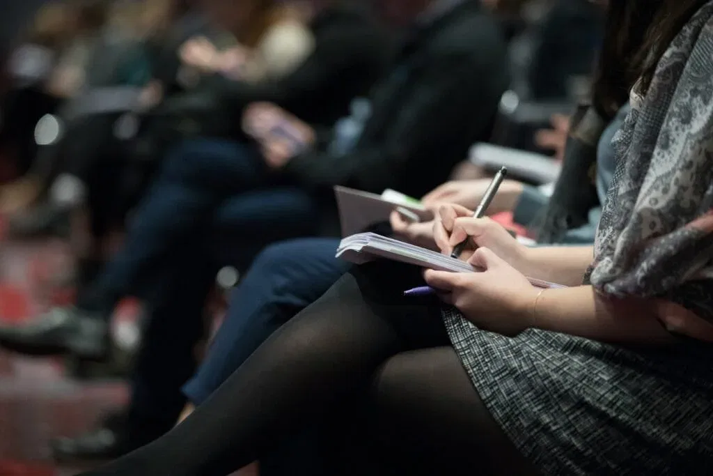 People seated in a room, attentively taking notes on notepads during an event or lecture.