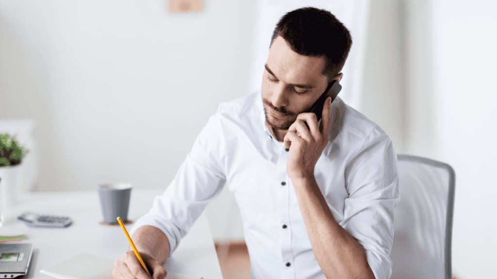 A man in a white shirt sits at a desk, talks on the phone, and writes in a notebook.