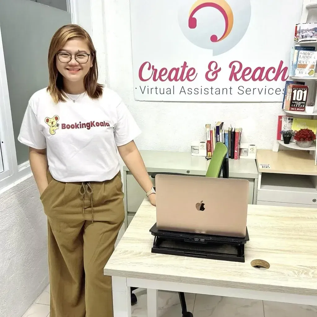 13 A person stands in an office wearing a BookingKoala shirt. Behind them is a desk with a laptop and a sign reading