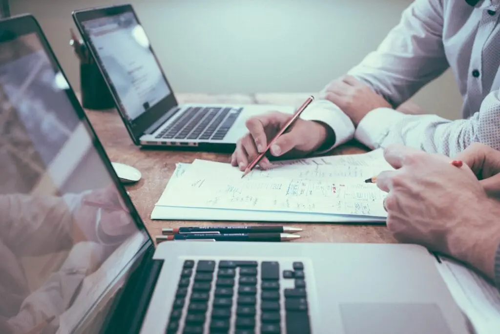 Two people work at a desk with laptops and papers, one pointing with a pencil.