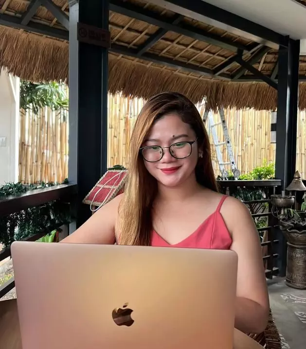 A person with long hair and glasses is working on a laptop at an outdoor cafe with bamboo decor.